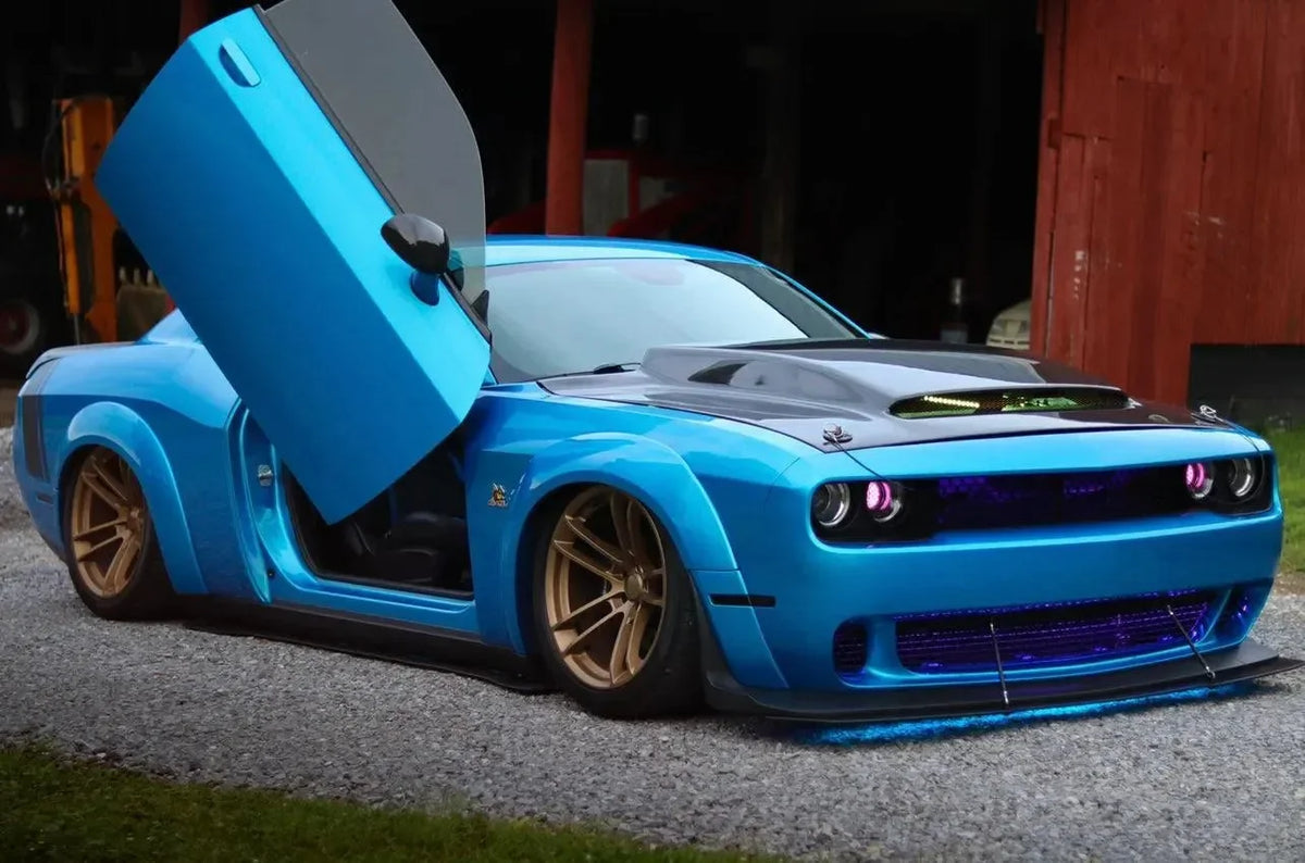 Blue Dodge Challenger with carbon fiber hood and custom gold wheels, parked outdoors near red barn