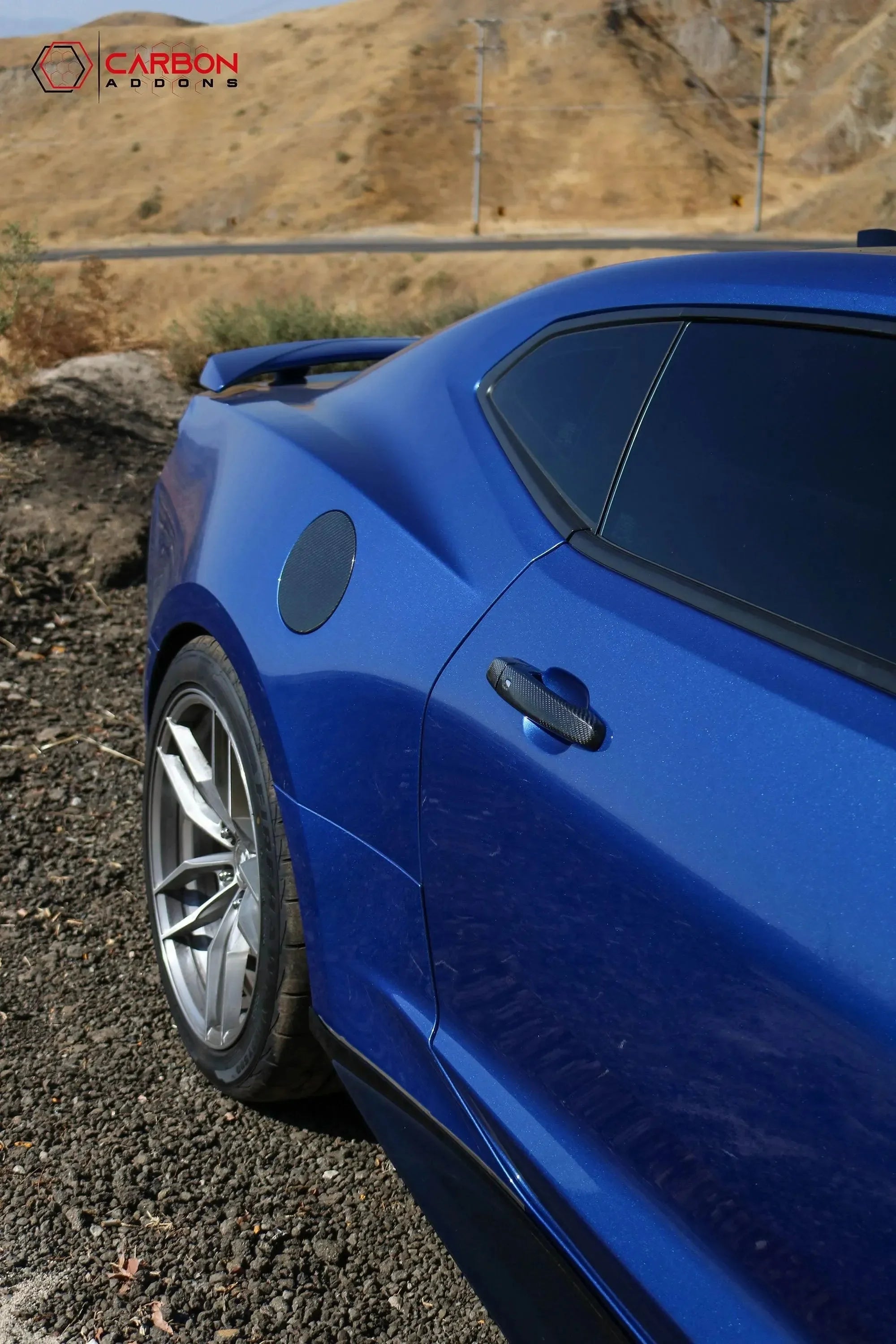 Blue sports car with carbon fiber door handle and side vent, parked on gravel near hills