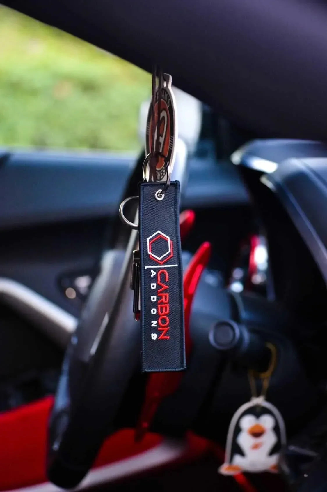 Carbon Addons keychain hanging in car interior with red accents and penguin charm